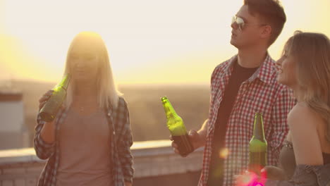 Three-girls-and-three-boys-enjoying-time-on-the-roof.-They-smile-and-communicate-with-each-other.-They-drink-beer-from-green-bottels-and-have-fun-in-the-plaid-shirts.